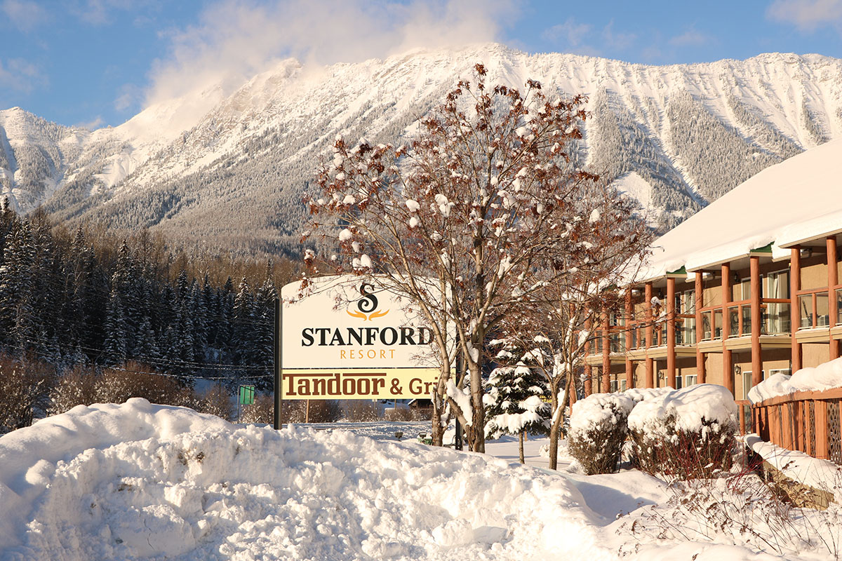 The Stanford Fernie Resort and Tandoor & Grill sign stands amongst snow covered trees, snow banks and the snow-capped Rocky Mountains in the background.